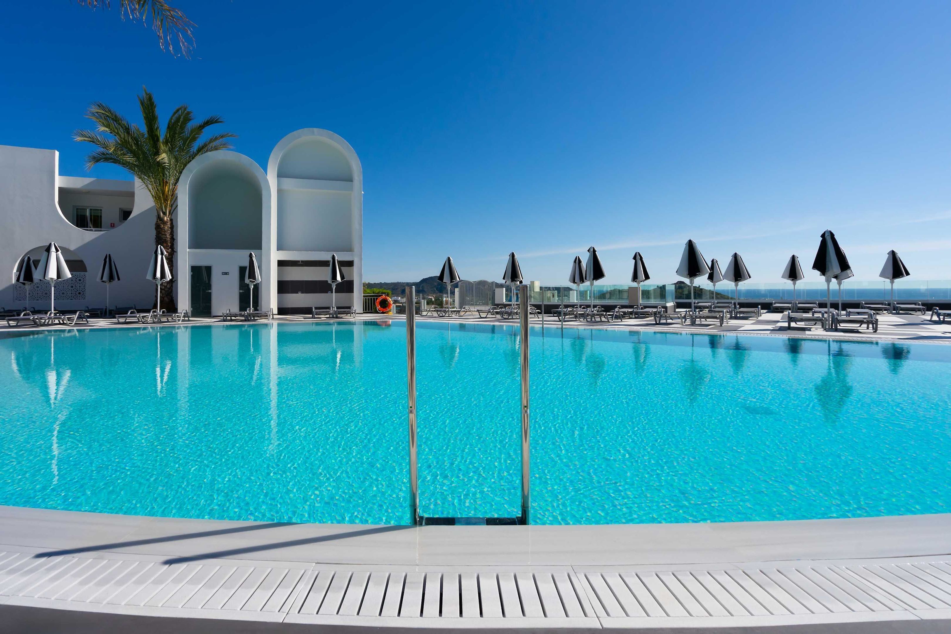 Une piscine d’un bleu éclatant, bordée de chaises longues et d’ombrelles, et entourée d’une architecture moderne et de palmiers.