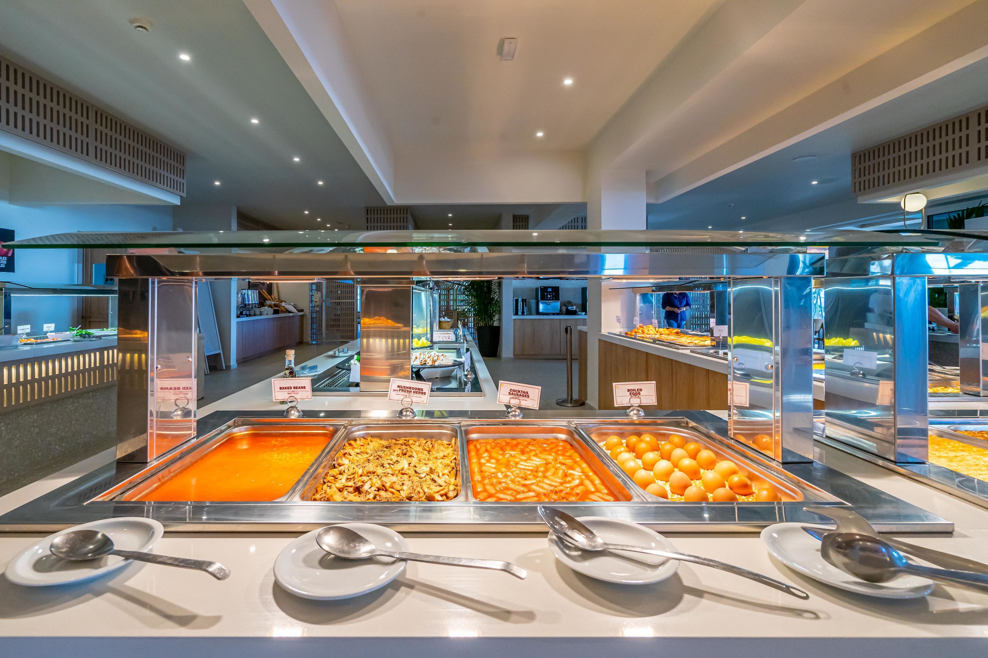 Uma área de buffet com várias estações de comida, incluindo opções como feijão cozido, massa e laranjas.