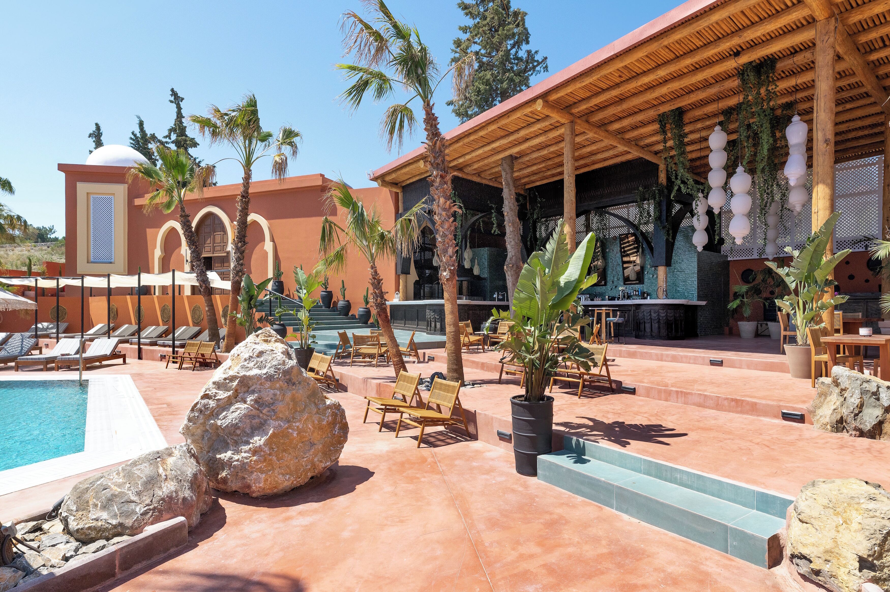 A pool area with lounge chairs, a bar, and a covered seating area with hanging lanterns.