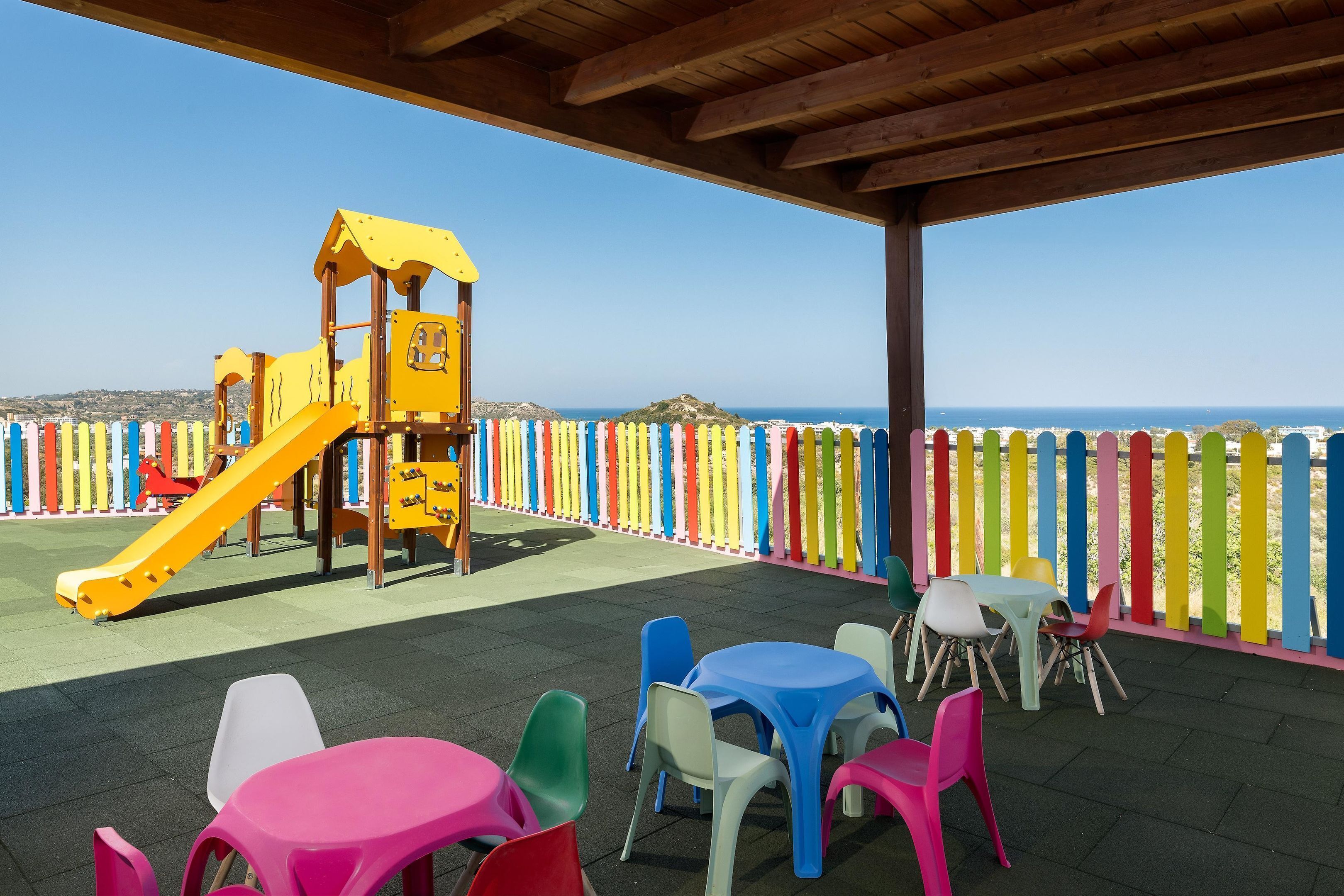 Un espace de jeux pour enfants avec un toboggan et une structure d’escalade, entouré de clôtures colorées, ainsi que des tables et des chaises d’extérieur.