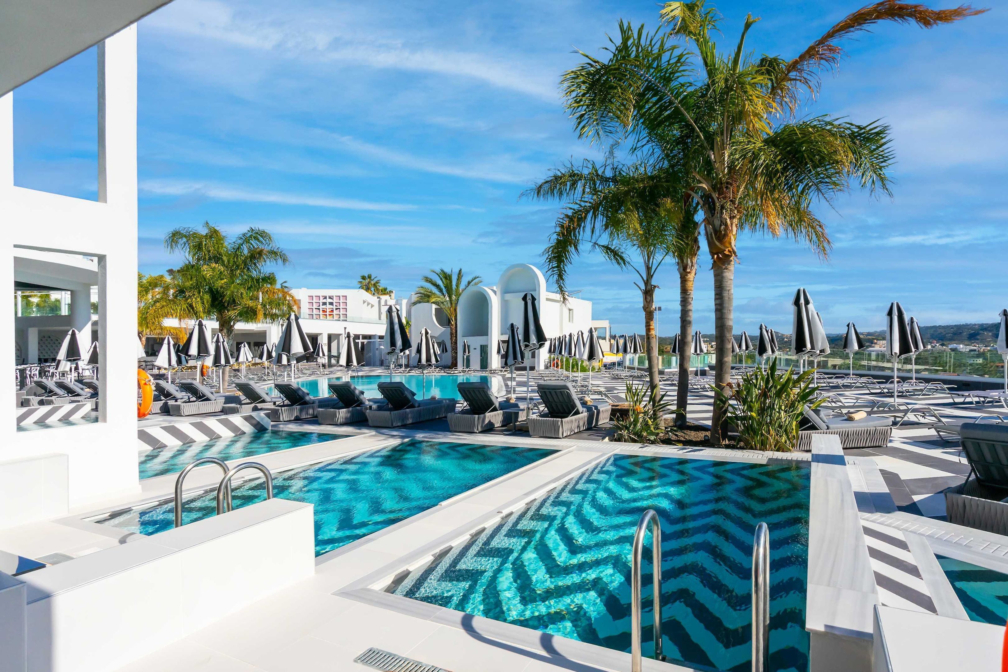 Un espace piscine doté de nombreuses chaises longues et de parasols, entouré de palmiers et bordé d’une architecture moderne.