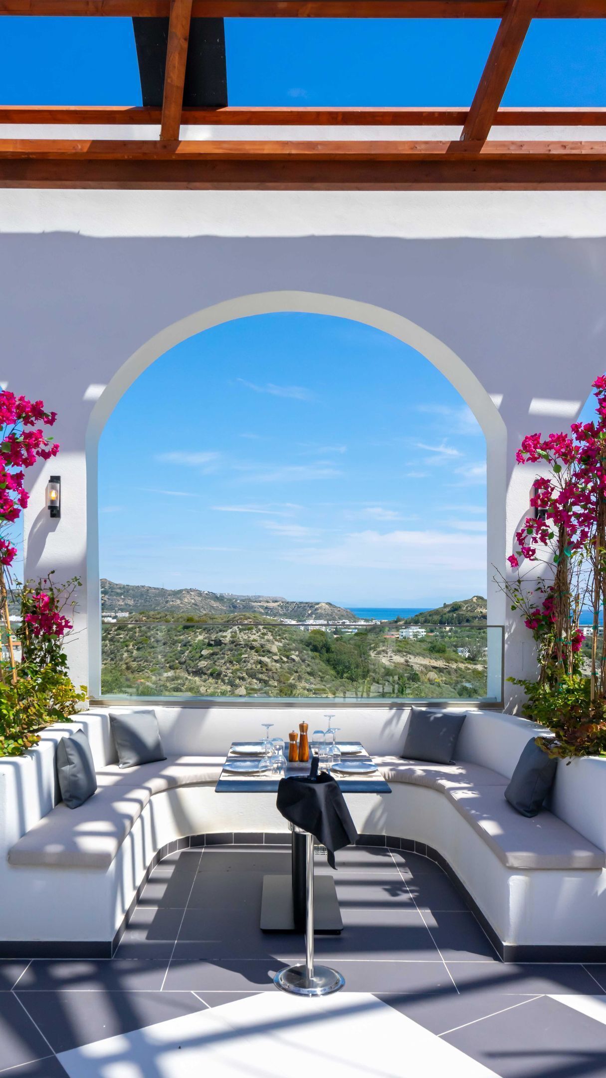 A modern outdoor seating area with a curved white sofa, a dining table set for two, and a clear view of a hilly landscape.