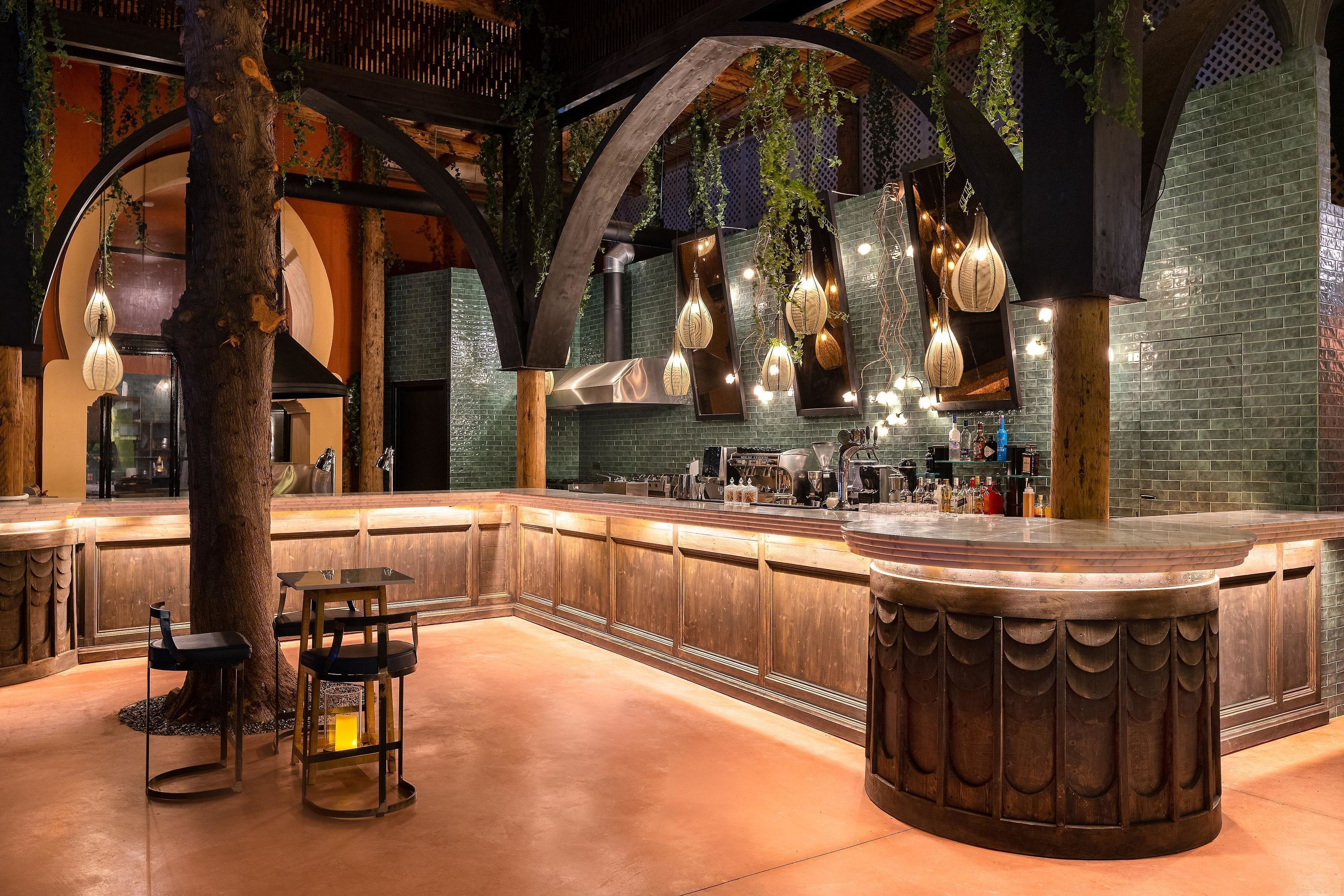 A bar area with a curved wooden counter, hanging pendant lights, and a large tree trunk centerpiece.