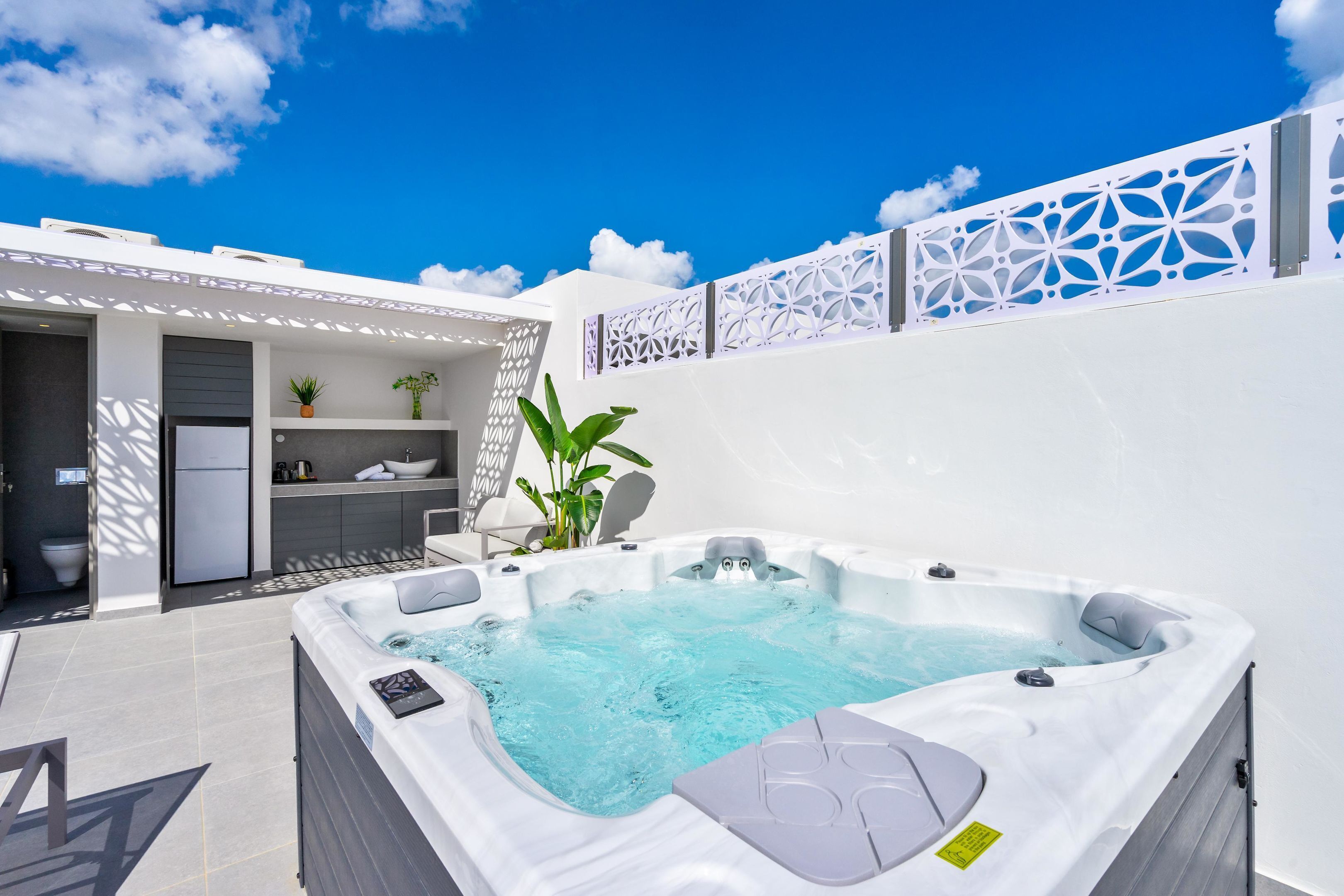 A modern outdoor hot tub area with a clear blue sky, a white wall with decorative cutouts, and a small indoor bathroom with a toilet and sink.