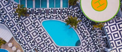 Vista aérea de un resort con una piscina central rodeada de zonas de descanso con diseños geométricos, otra piscina separada y un área circular con un logotipo.