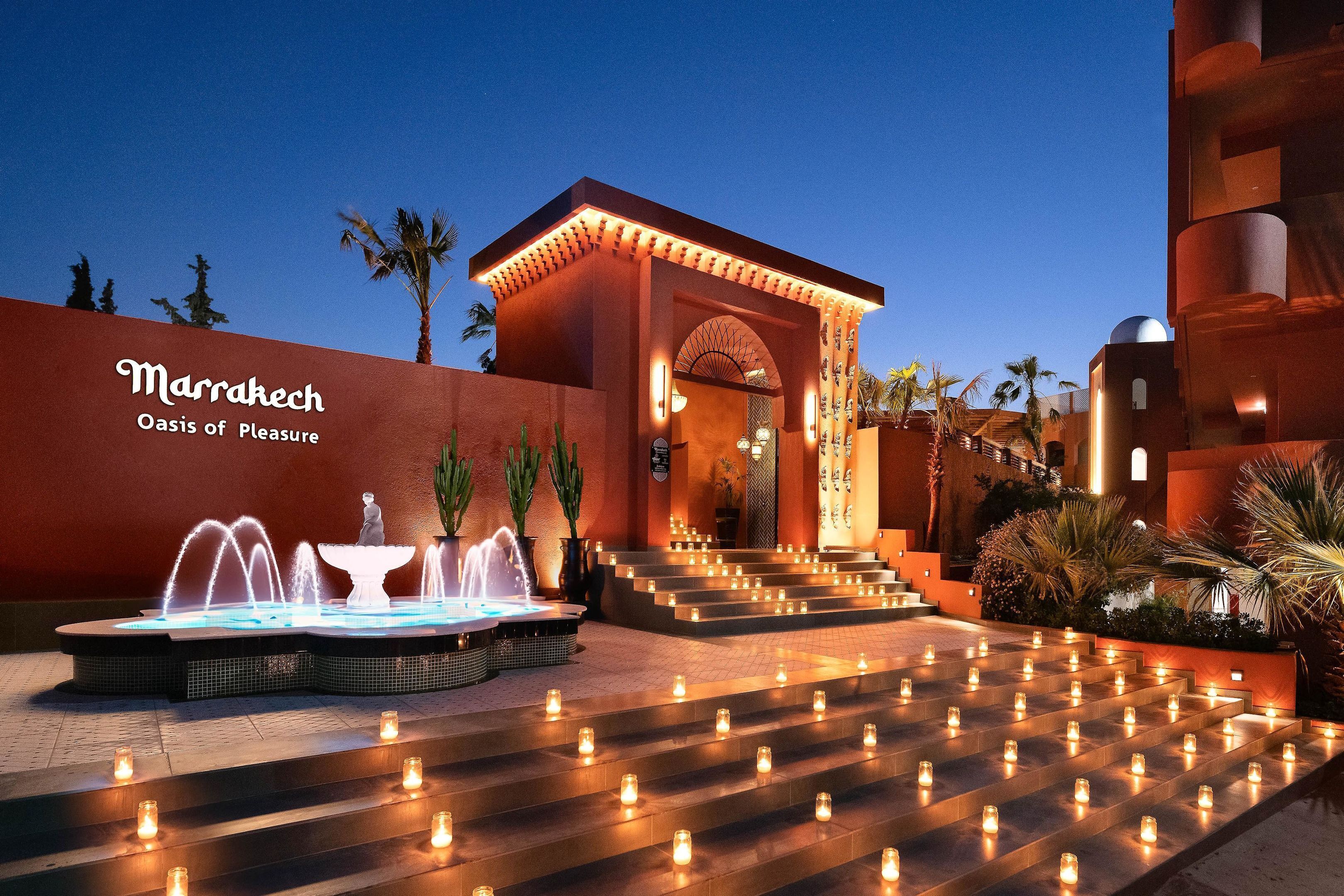 A hotel entrance with a fountain, steps adorned with candles, and a sign reading "Marrakech Oasis of Pleasure."