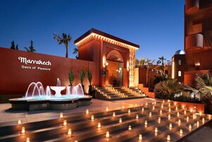 Entrada de un hotel con una fuente, escalones decorados con velas y un letrero que dice 'Marrakech Oasis of Pleasure'.