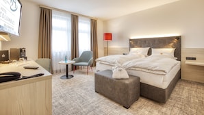 A hotel room with a bed, a desk, a chair, a small table, and a lamp. - Trans World Hotel Columbus (Seligenstadt)