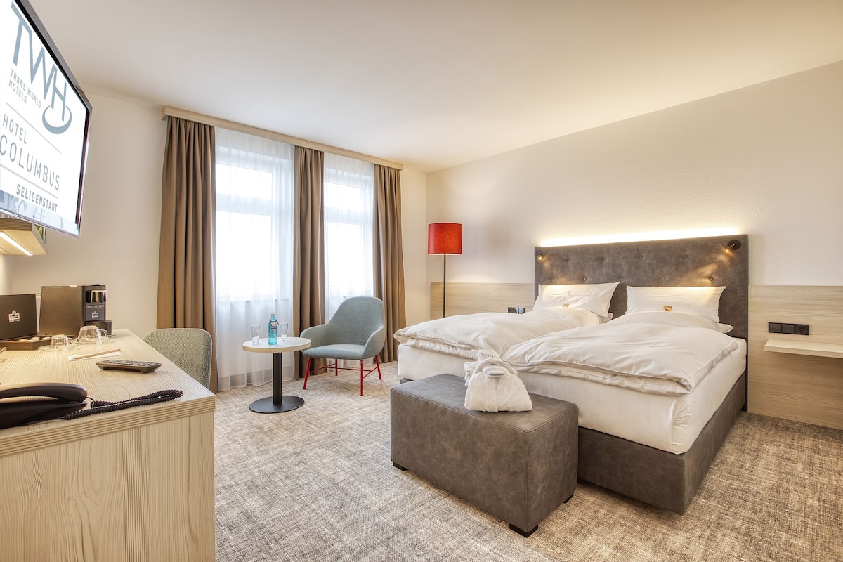 A hotel room with a bed, a desk, a chair, a small table, and a lamp.