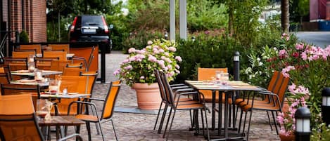 An outdoor cafe with tables and chairs, a potted plant, and a brick building in the background.