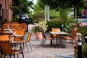 An outdoor cafe with tables and chairs, a potted plant, and a brick building in the background. - Trans World Hotel Columbus (Seligenstadt)