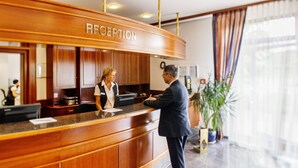 A reception area with a wooden reception desk, a receptionist, and a guest. - Trans World Hotel Columbus (Seligenstadt)