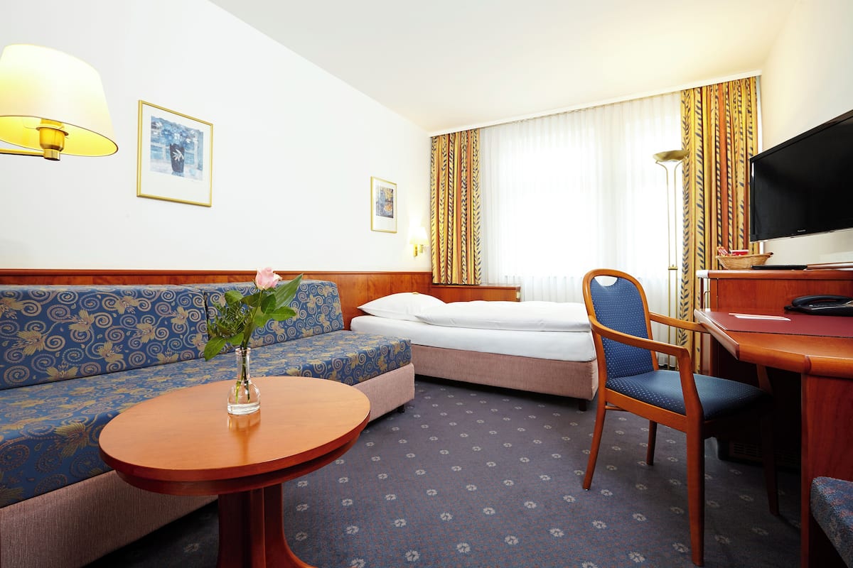 A hotel room with a sofa, a bed, a desk with a television, and a small table with a vase.