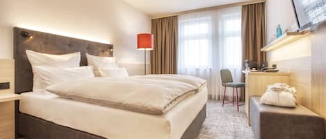 Une chambre d’hôtel avec un grand lit, un bureau avec une chaise, un petit pouf et un téléviseur fixé au mur.