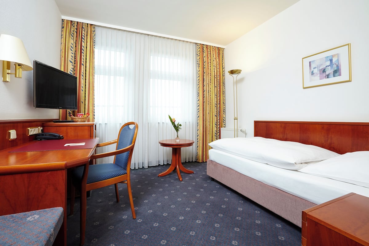 A hotel room with a bed, a desk with a chair, a television, a small table with a plant, and curtains.