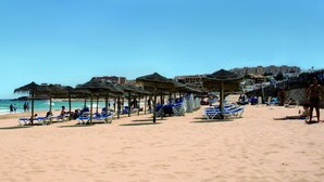 Une plage avec des transats et des parasols, un ciel dégagé et des bâtiments en arrière-plan.