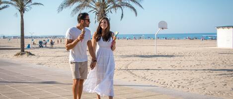 Una pareja caminando por un paseo marítimo con palmeras y un aro de baloncesto al fondo.