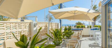An outdoor seating area with white chairs and large beige umbrellas, overlooking a beach with palm trees and a clear blue sky.