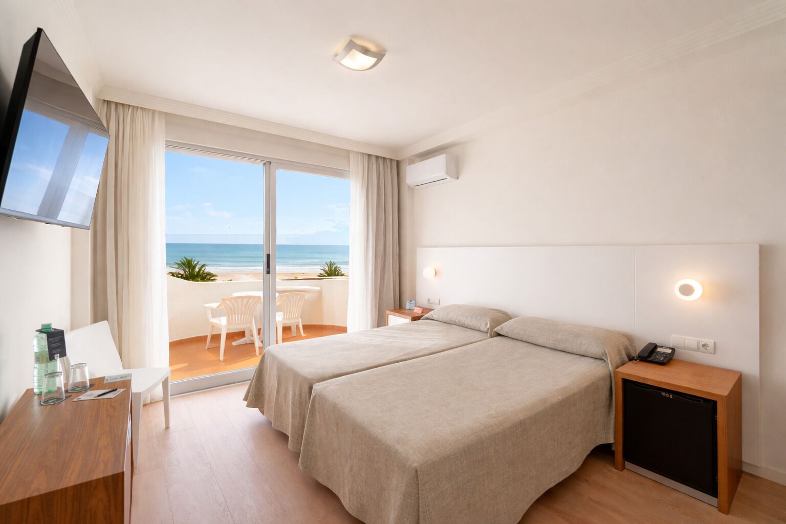 A hotel room with a bed, a desk, a chair, a TV, and a view of the beach.