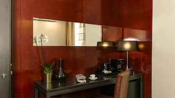 A hotel room with a wooden wall, a mirror, a desk with a lamp, a chair, a small potted plant, a vase, a coffee maker, and a newspaper.