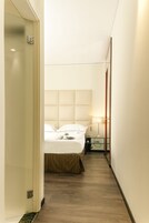 Un corridoio d'albergo stretto con un letto in fondo, un comodino con una lampada, e una porta a sinistra.