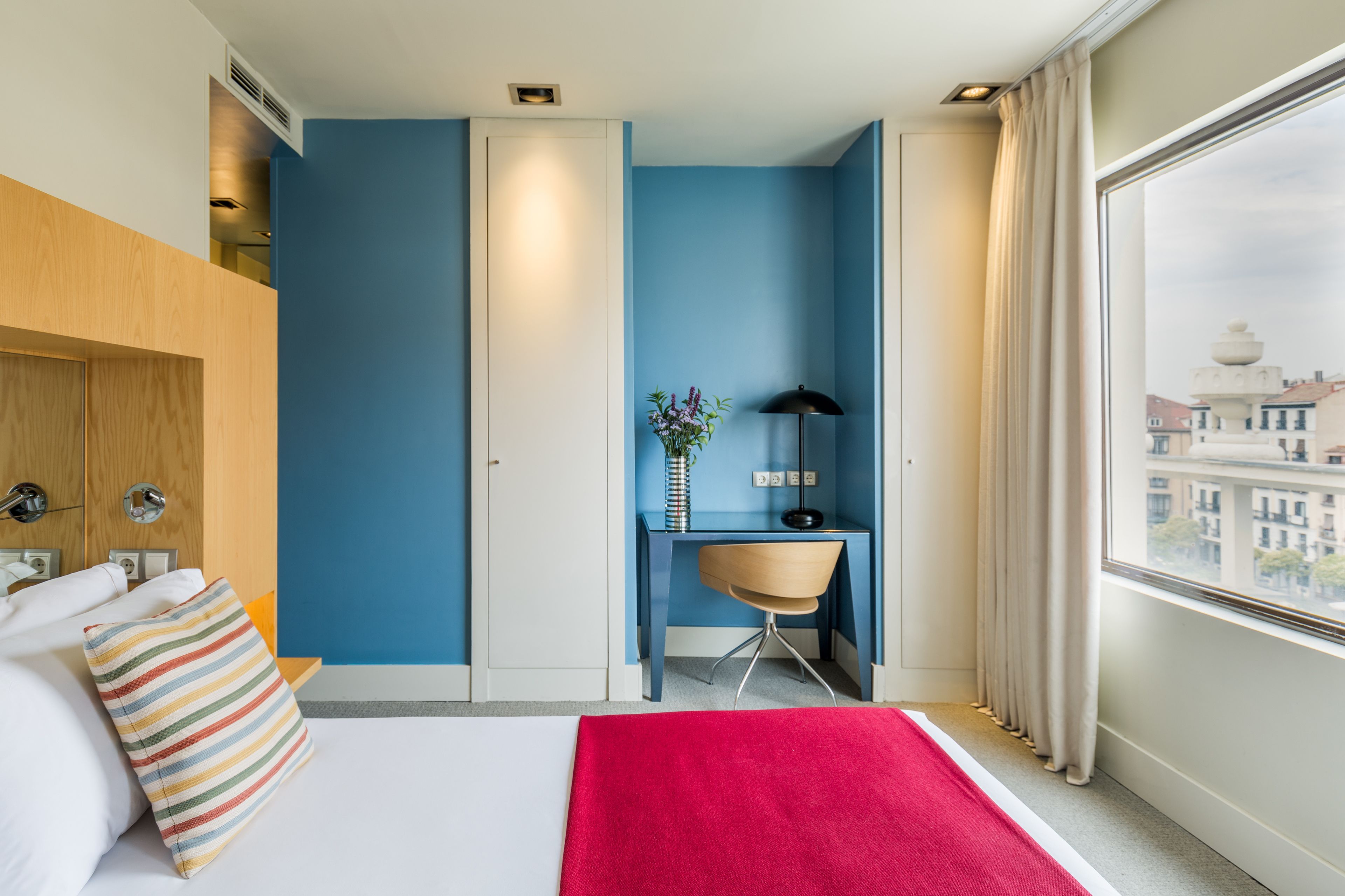 Habitación de hotel con cama, escritorio, silla, ventana con cortinas y vista a edificios.