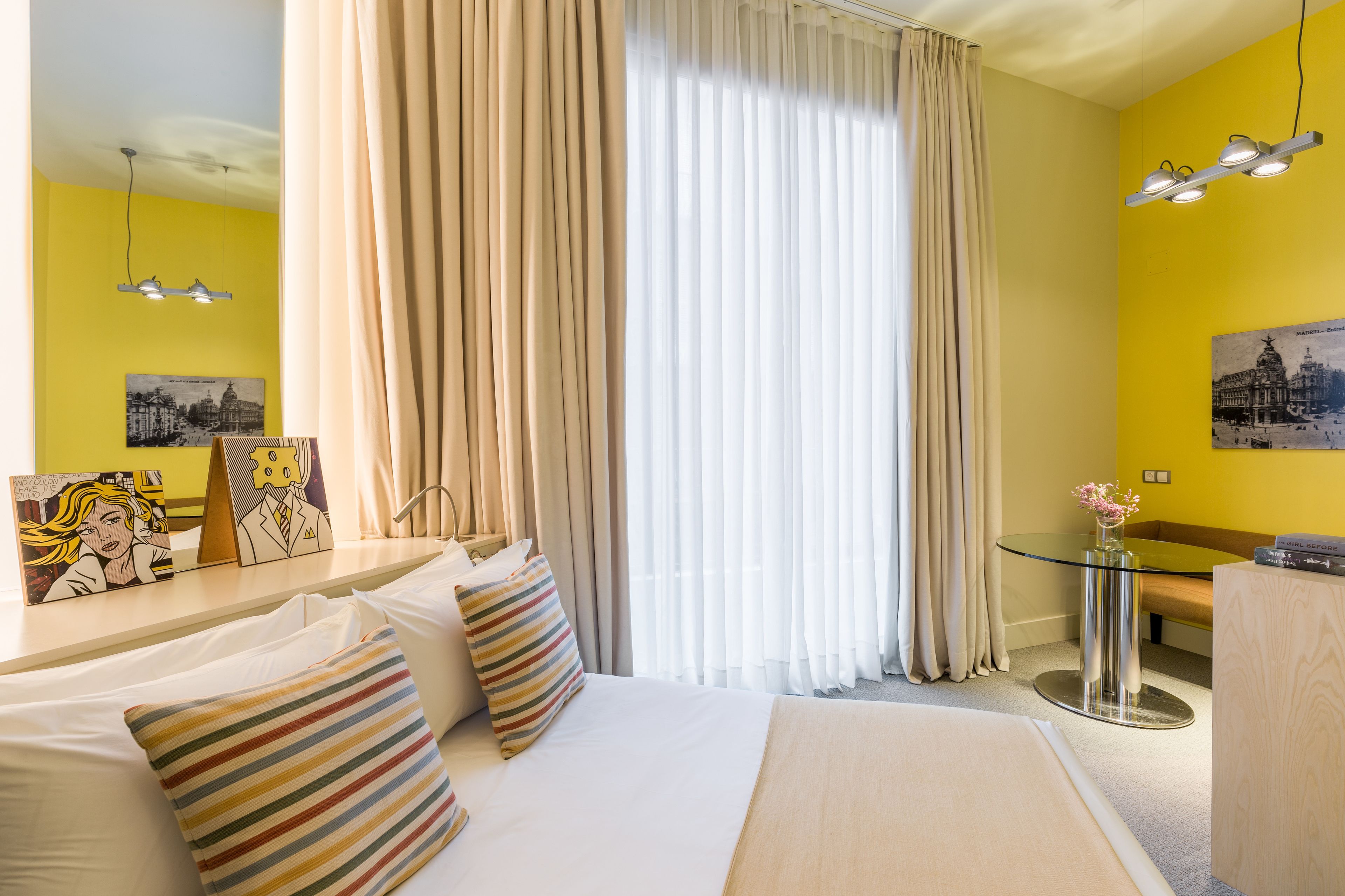 A hotel room with a bed, a bedside table, a glass table, and curtains.