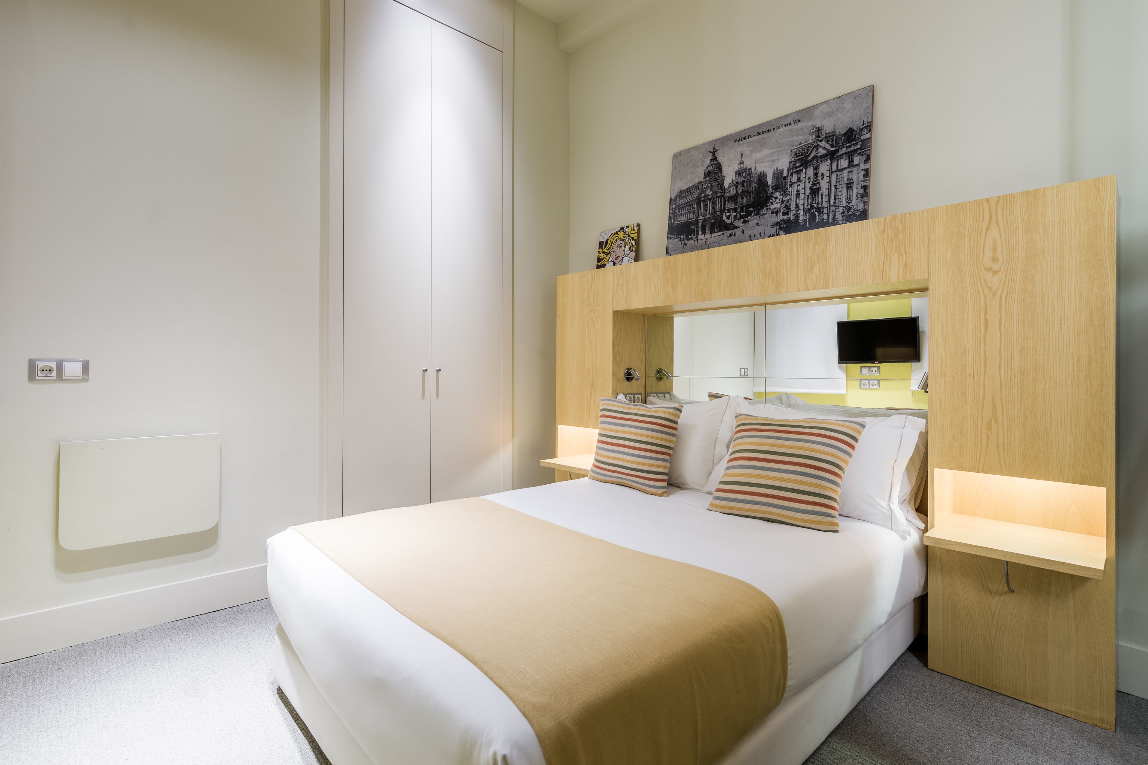 Habitación de hotel ordenada con cama, mesitas de noche, armario y televisor montado en la pared.