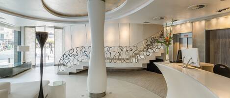 Un área de recepción moderna con un mostrador curvo, una columna grande y una escalera adornada con arte abstracto en la pared.