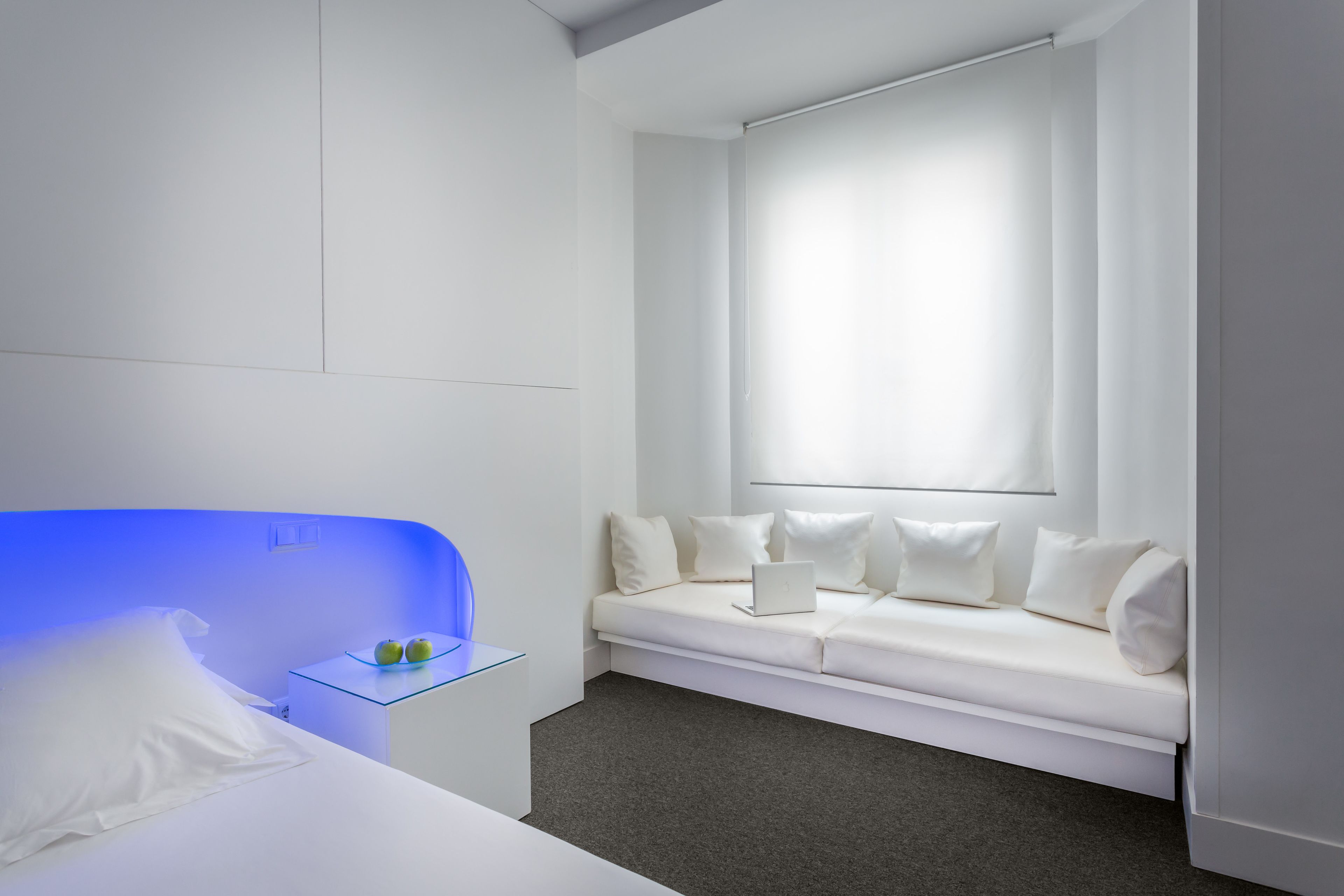 A modern hotel room with a white bed, a blue-lit headboard, a white sofa with a laptop, and a large window with curtains.