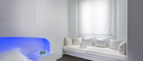 Una habitación de hotel moderna con una cama blanca, un cabecero iluminado en azul, un sofá blanco con una computadora portátil y un ventanal con cortinas.