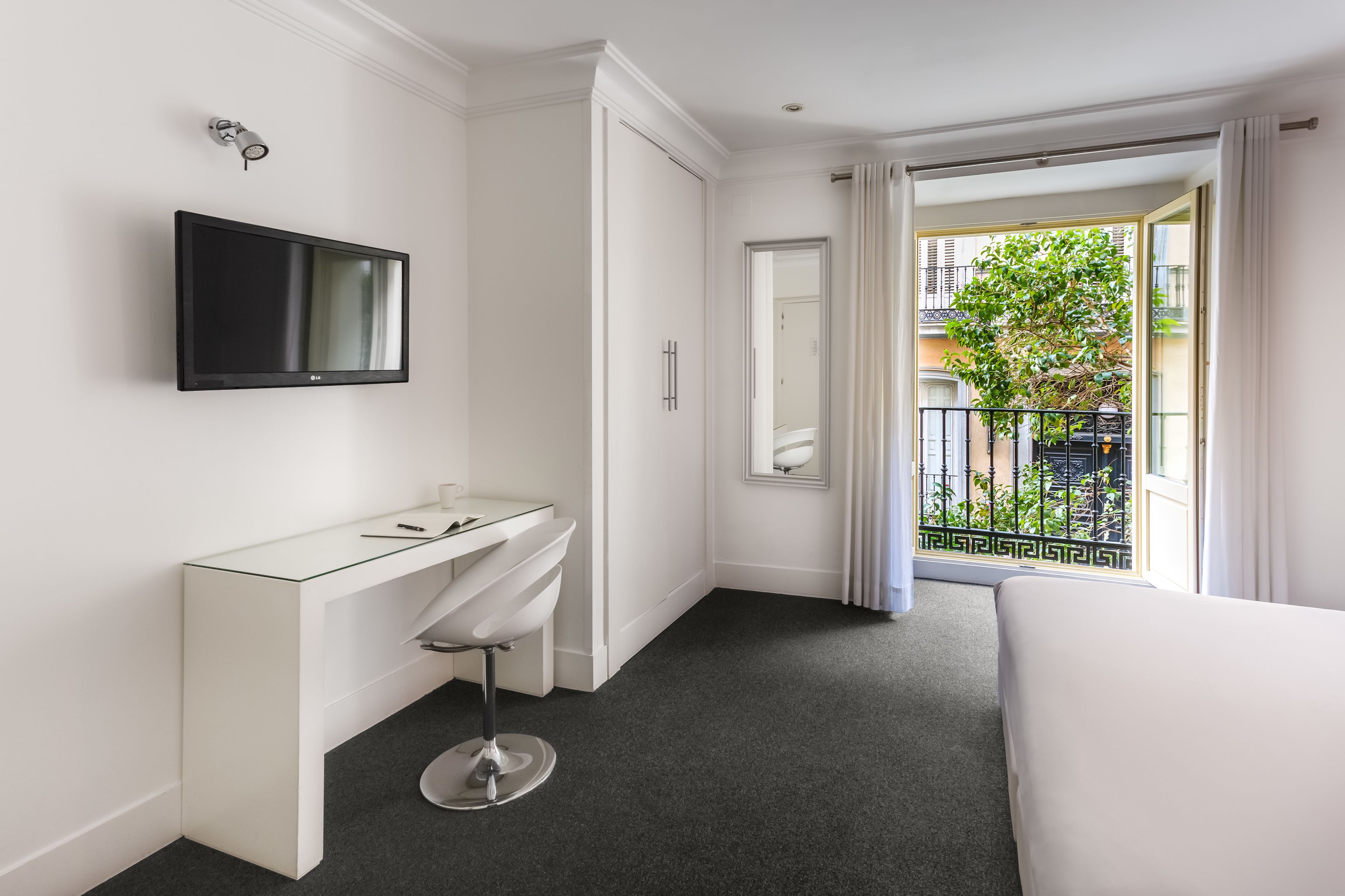 A modern hotel room with a large bed, a white vanity with a mirror, a flat-screen TV mounted on the wall, and a balcony with a view of the city.