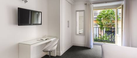 A modern hotel room with a large bed, a white vanity with a mirror, a flat-screen TV mounted on the wall, and a balcony with a view of the city.