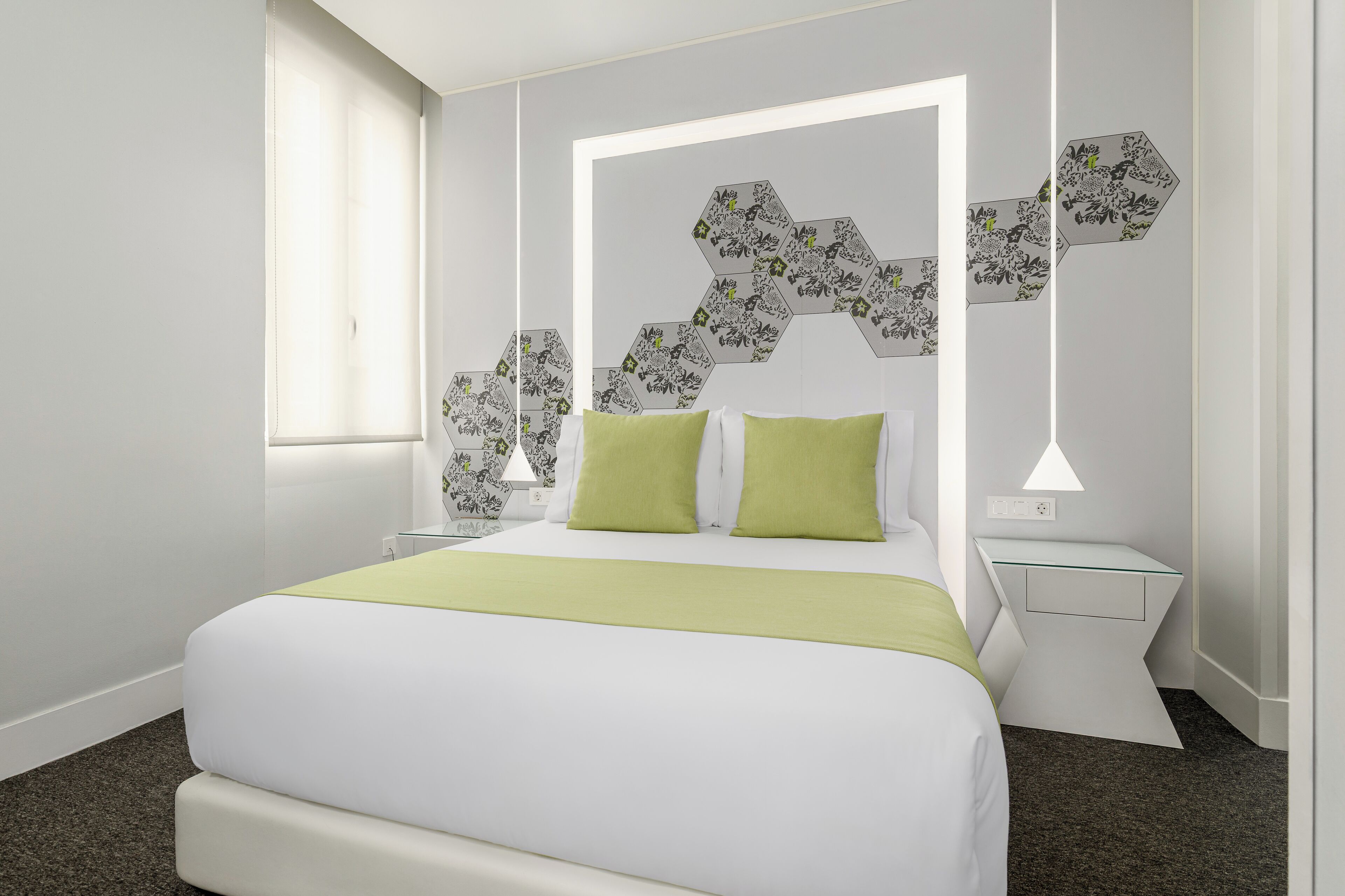 A modern hotel room with a large bed, a nightstand, a wall-mounted lamp, and a window with blinds.