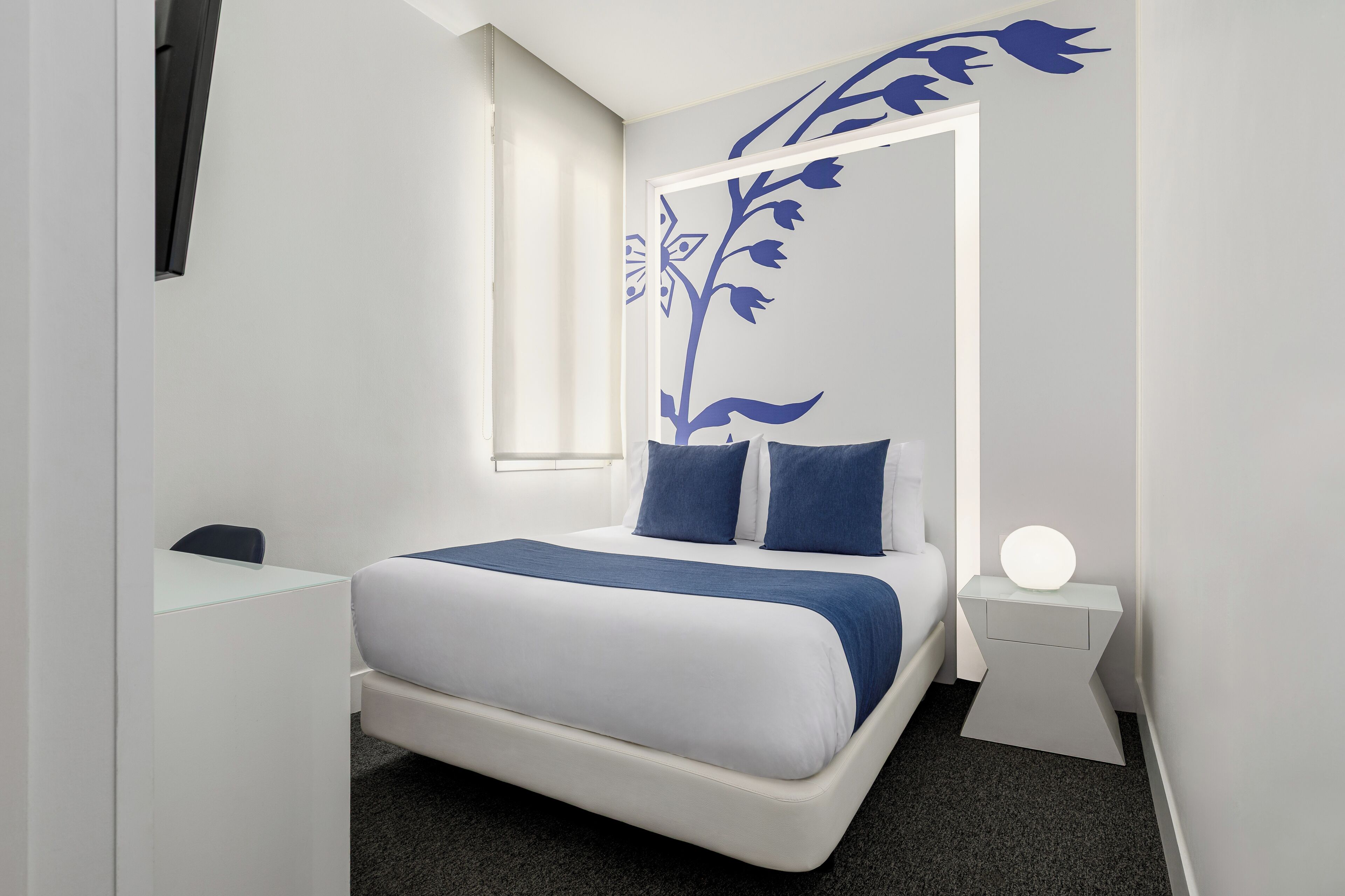 Habitació d'hotel moderna amb un llit, una escriptori i una tauleta de nit amb una làmpada.