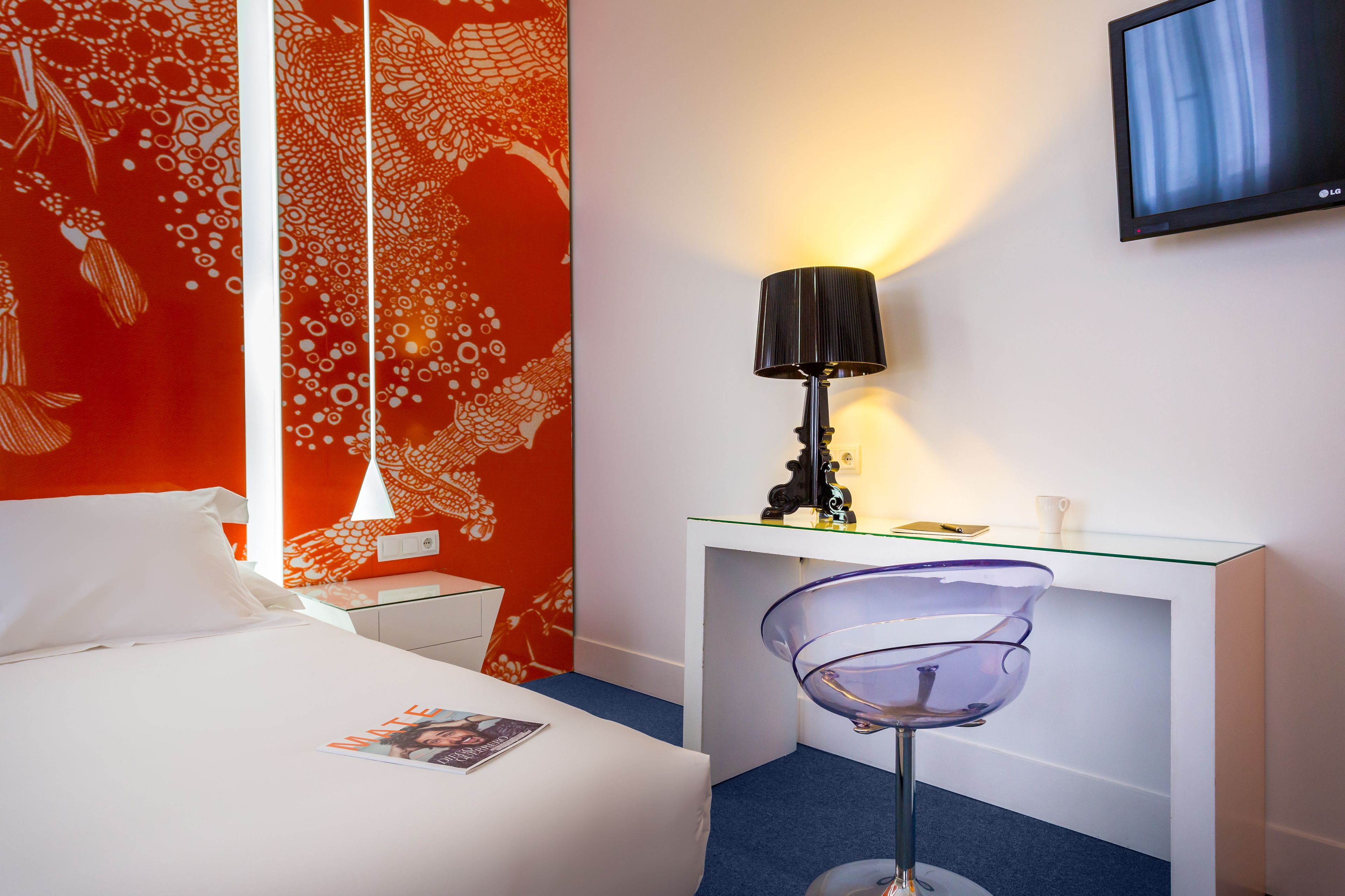 A hotel room with a bed, a desk with a lamp, a TV, and a chair.