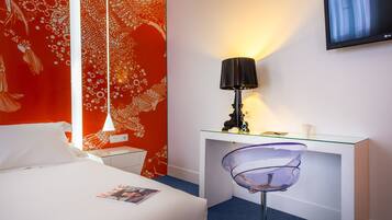 A hotel room with a bed, a desk with a lamp, a TV, and a chair.