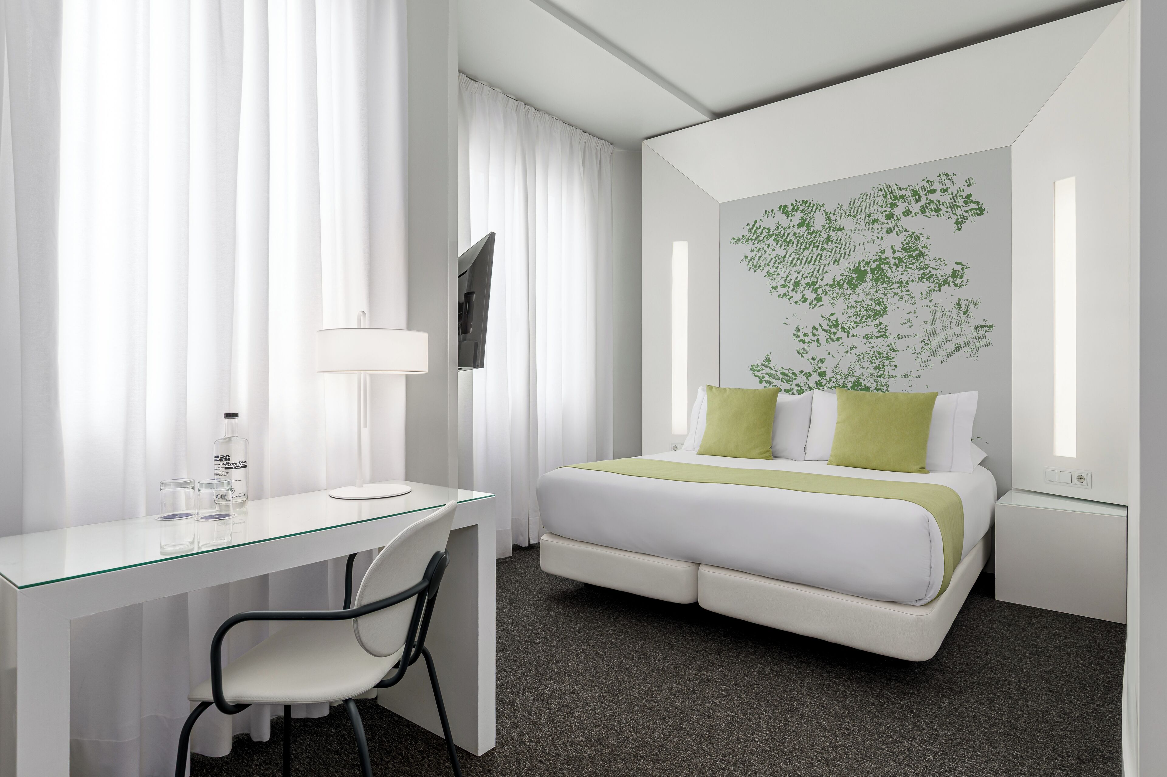 Una habitación de hotel moderna con un escritorio de vidrio, una cama con ropa de cama verde y blanca, un panel de arte mural con plantas verdes y un televisor de pantalla plana en la pared.