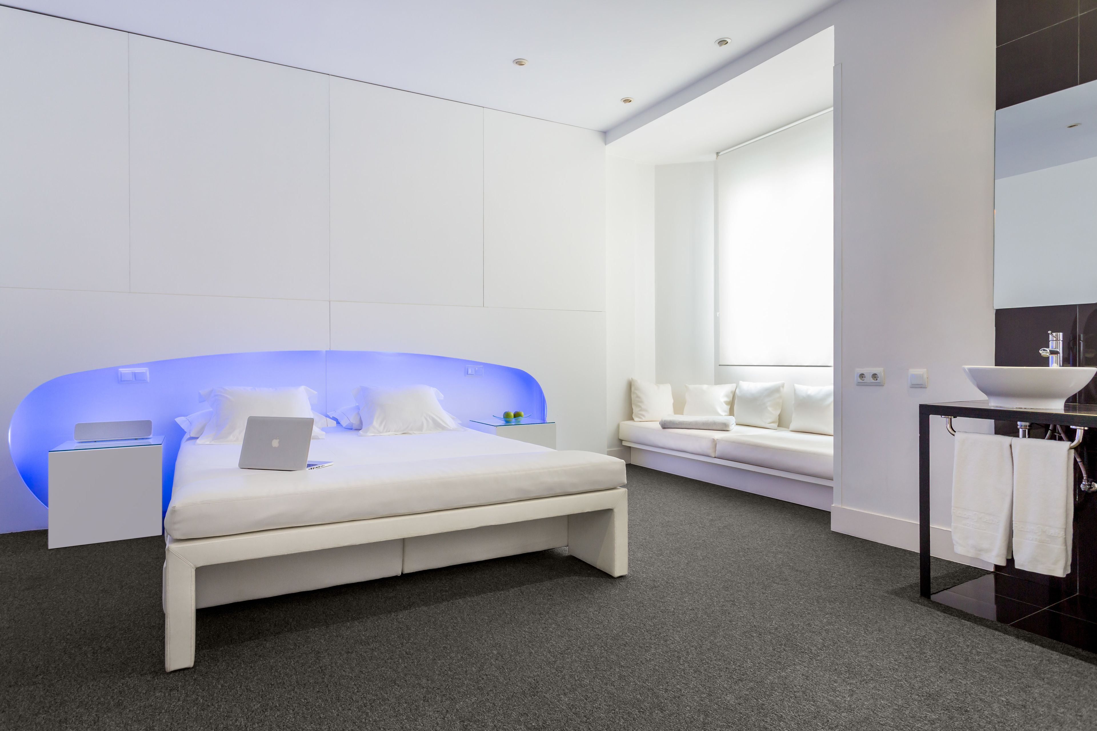 Ein modernes Schlafzimmer mit Bett, Nachttisch mit Laptop, Nachttischlampe, Fenster und einem Badezimmer mit Waschbecken und Handtuchhalter.