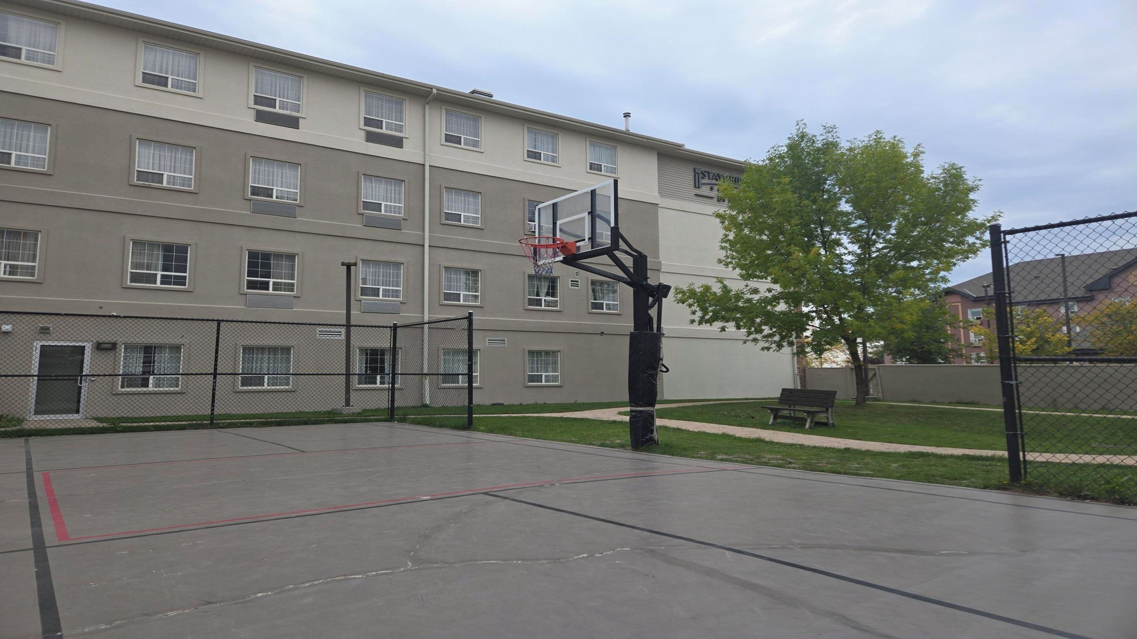 An outdoor basketball court with a hoop, surrounded by a chain-link fence, in front of a multi-story building with windows.