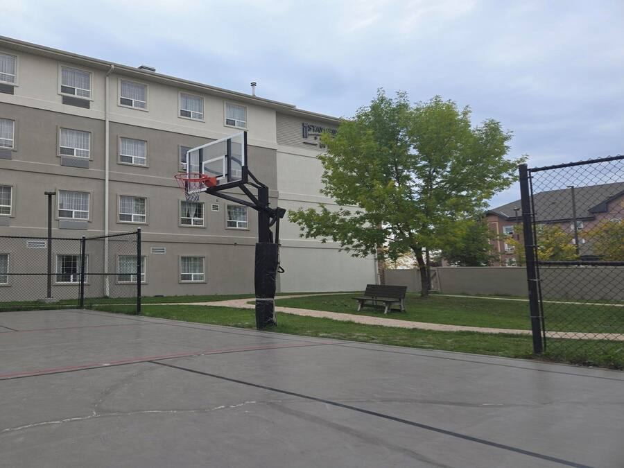 An outdoor basketball court with a hoop, surrounded by a chain-link fence, in front of a multi-story building with windows.