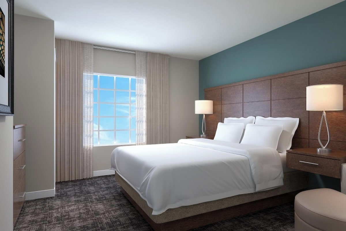 Una habitación de hotel con una cama grande, dos lámparas de noche, una silla y un ventanal con cortinas.