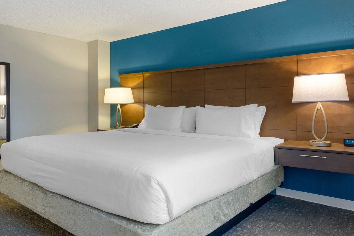 A hotel room with a large bed, two bedside lamps, a nightstand with a clock, and a mirror on the wall.