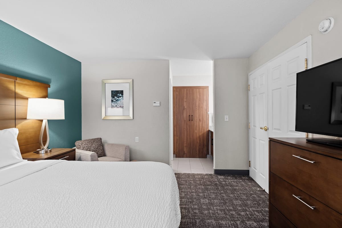 A hotel room with a bed, a TV, a desk with a lamp, a chair, a sofa, a framed picture on the wall, and a wardrobe.