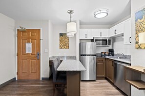 A modern kitchen with a stainless steel refrigerator, microwave, and dishwasher, wooden cabinets, a marble countertop, and a bench. - Staybridge Suites Calgary Airport by IHG (Calgary)