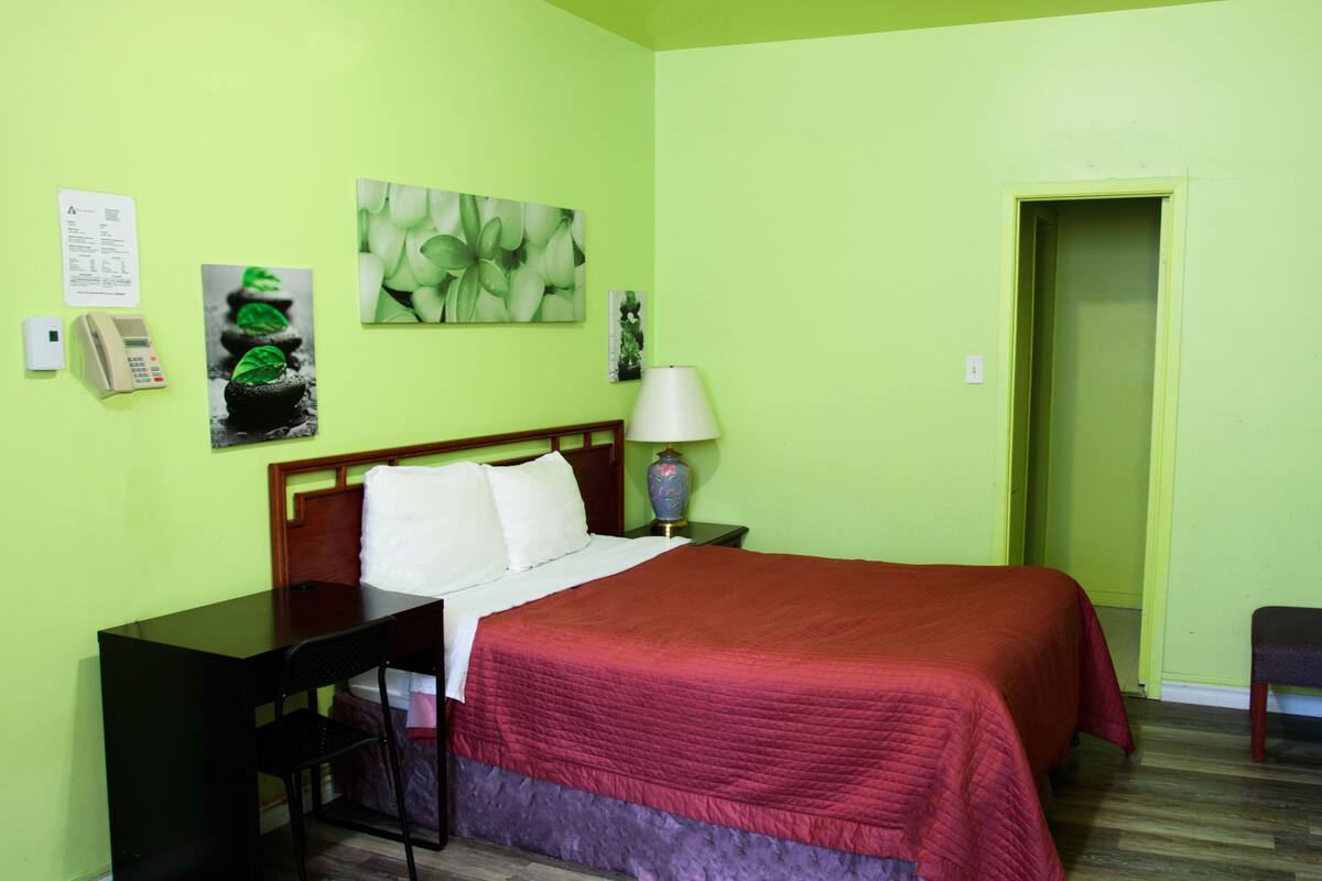 A hotel room with a bed, a desk, a chair, a lamp, and a door.