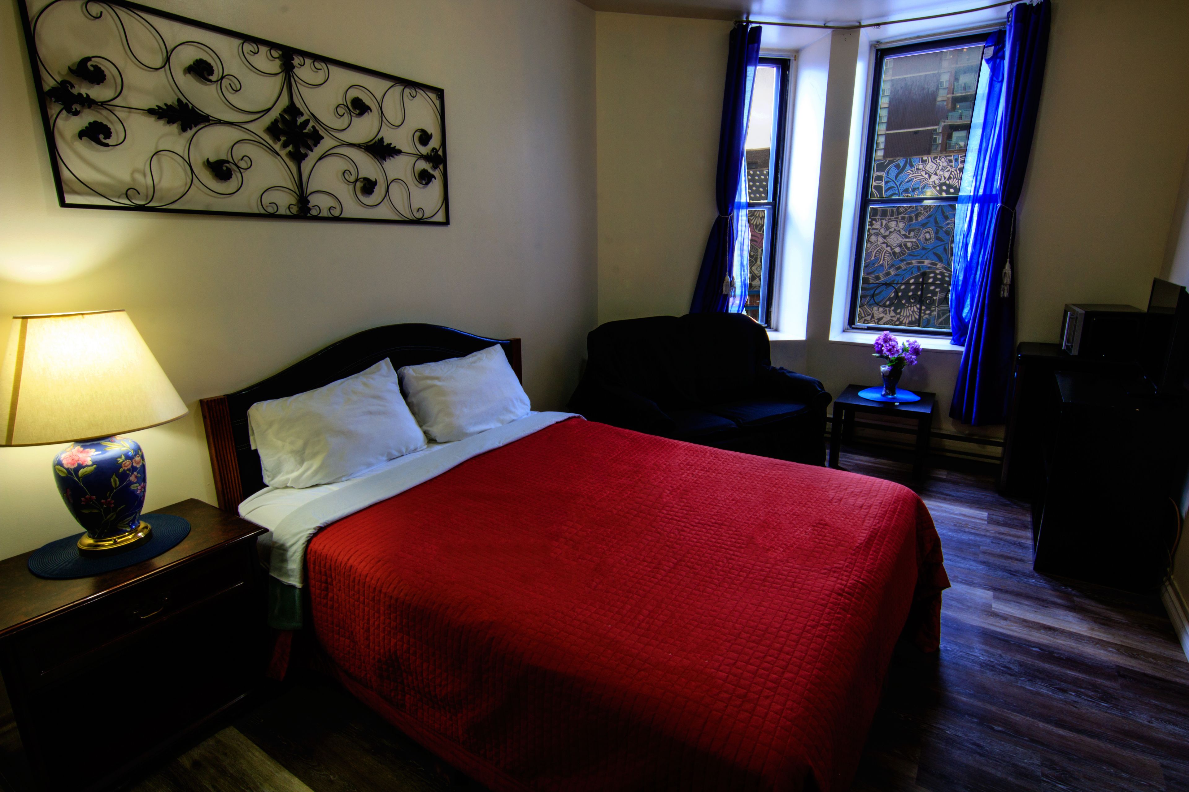 A hotel room with a bed, a nightstand with a lamp, a chair, a small table with a vase of flowers, and a window with curtains.