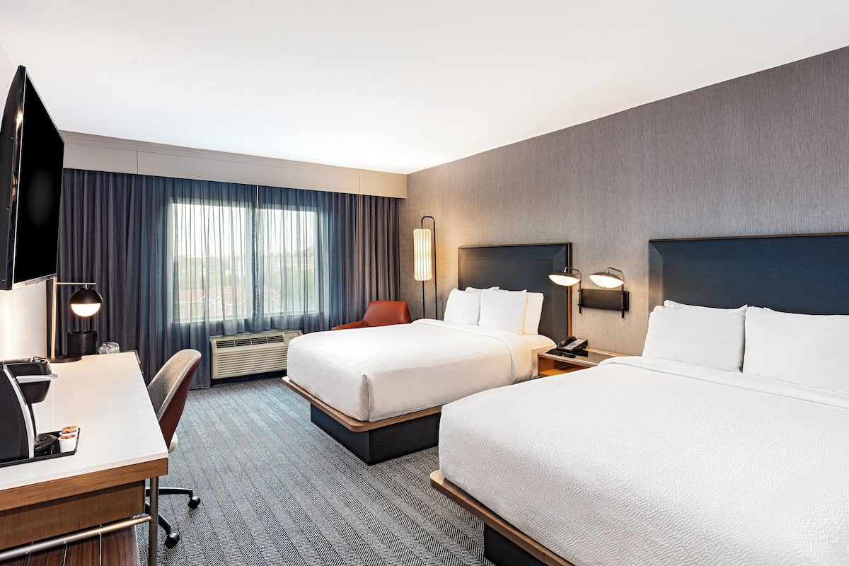 A hotel room with two beds, a desk, a chair, a TV, and a window with curtains.