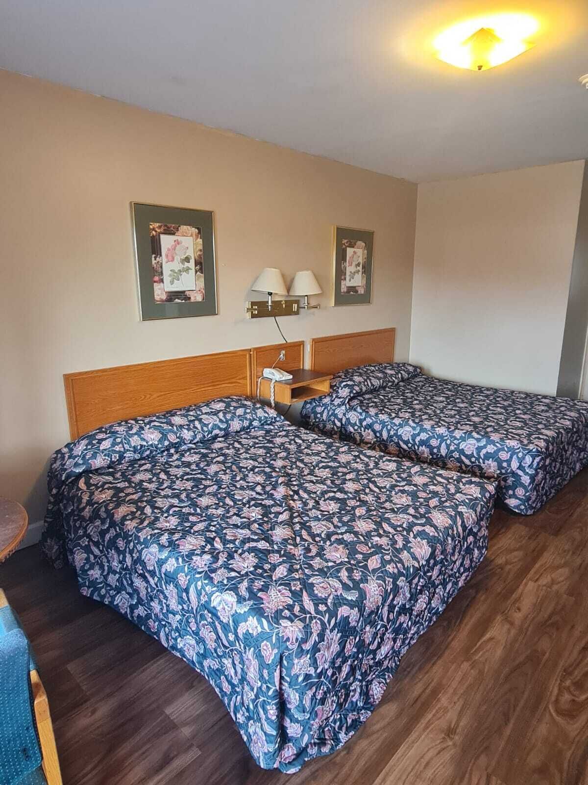 A room with two beds, a wooden headboard, a nightstand with a lamp, and framed pictures on the wall.