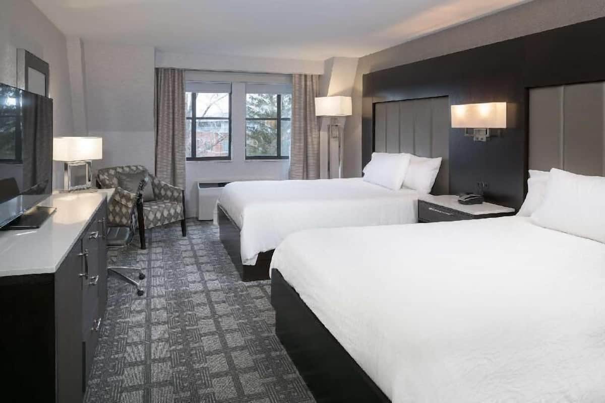 Ein Hotelzimmer mit zwei Betten, einem Schreibtisch, einem Stuhl, einem Fernseher und einem Fenster mit Vorhängen.